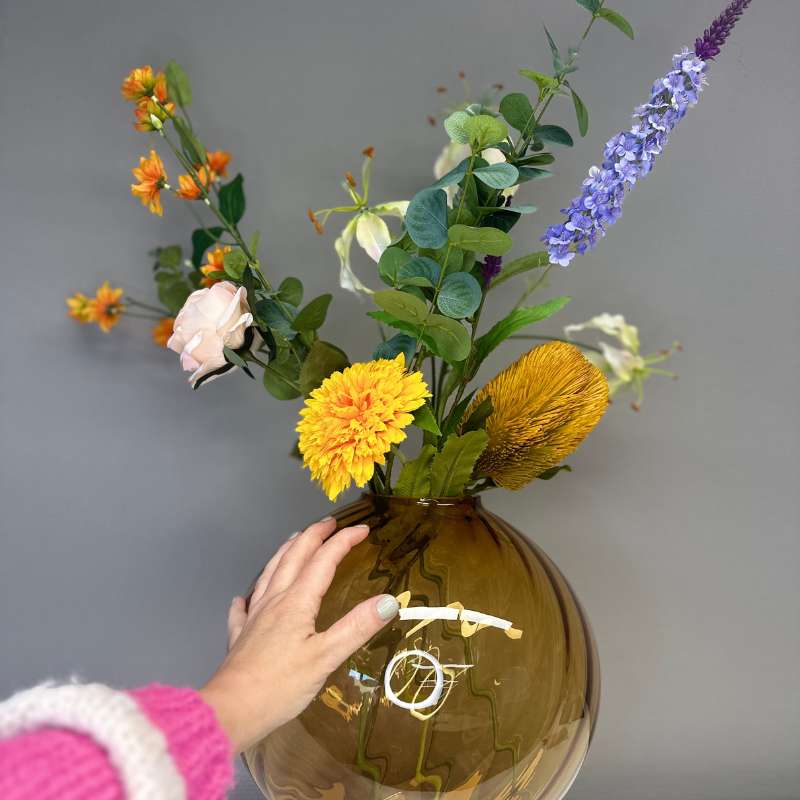 Boeket BeeBee met zijden bloemen in oranje en paars tinten met een hand op vaas. Vaas Fridio mondgeblazen collectie BloomFever